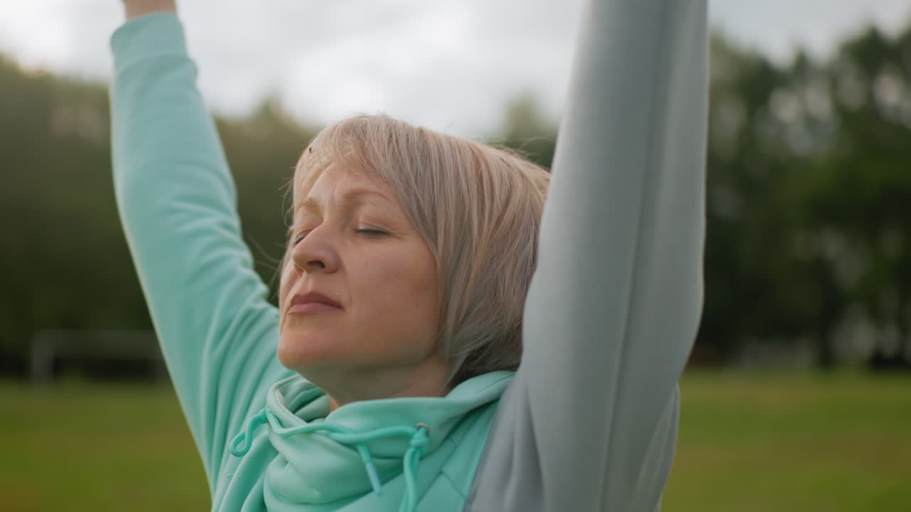Fair skinned lady with short blond hair wearing blue hoodie practicing controlled breath exercise outdoors eyes closed arms stretched in peaceful green park under soft daylight enjoying tranquility