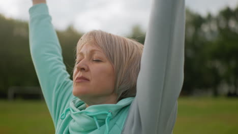 fair skinned lady with short blond hair wearing blue hoodie practicing controlled breath exercise outdoors eyes closed arms stretched in peaceful green park under soft daylight enjoying tranquility