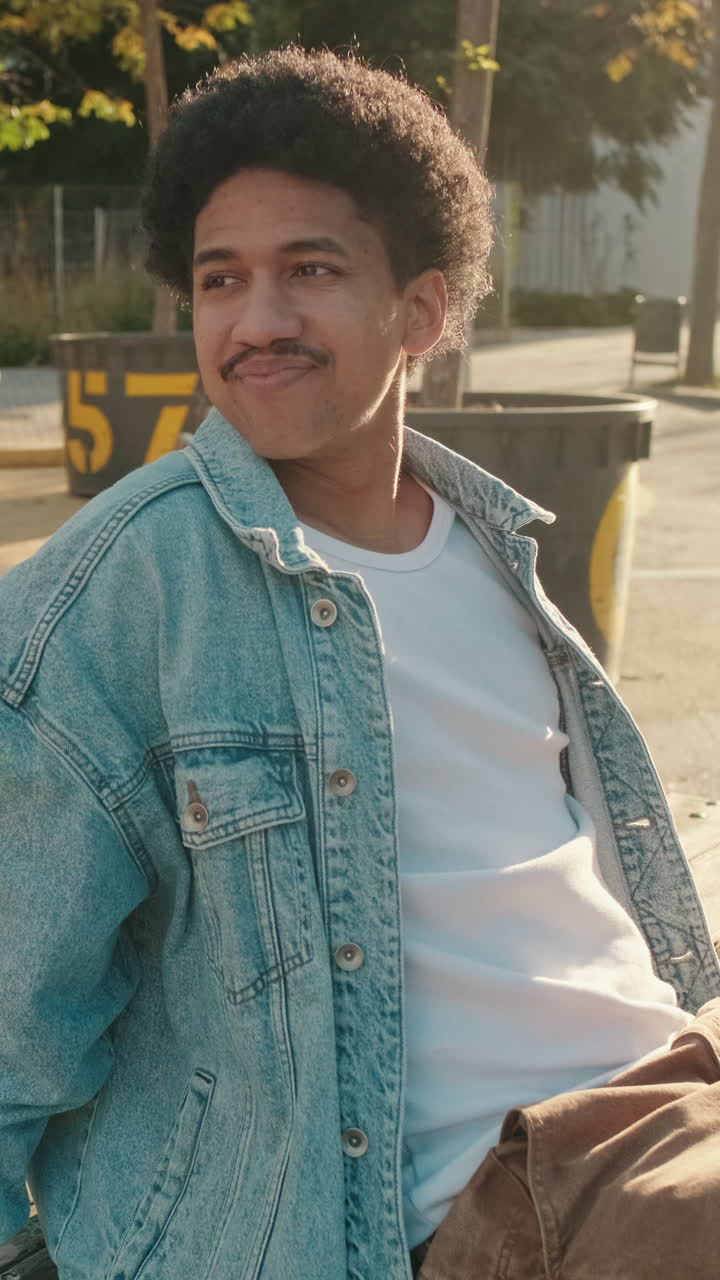 Jovem com cabelo cacheado e bigode sentado ao ar livre e sorrindo