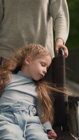 woman pushes wheelchair with little daughter walking in park on sunny spring day. girl with spinal paralysis sleeps in medical equipment closeup