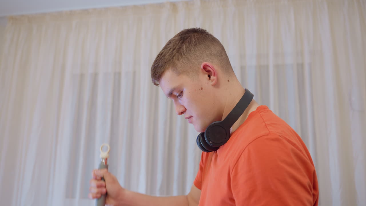Young boy in orange shirt with headphones around neck dipping mop handle into water bucket and lifting it out while focusing on home cleaning routine, showing responsibility, and hygiene process