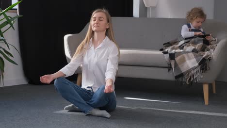 young mother doing yoga sitting in lotus position at home in the middle of the living room young mother doing yoga sitting in lotus position at home in the middle of the living room a child plays mobile games meditation at home
