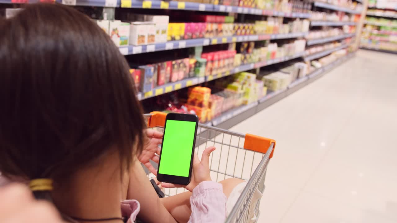 스마트폰의 녹색 화면을 가진 쇼핑 카트에 앉아있는 갈색 소녀의 뒷면 사진과 제품과 함께 선반 사이를 는 카트를 밀고있는 사람이 있습니다.