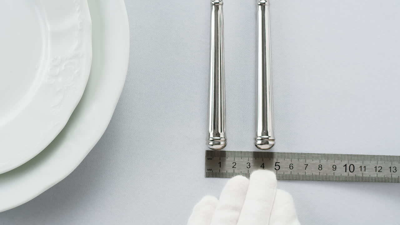Minimalist table setting with white plate, fork, and knife arranged neatly, and a gloved hand adjusting cutlery.