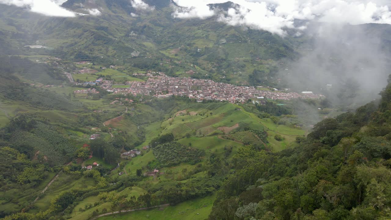 Drone ascends hills through clouds to frame Jardin town center, surrounded by green mountains in Colombia