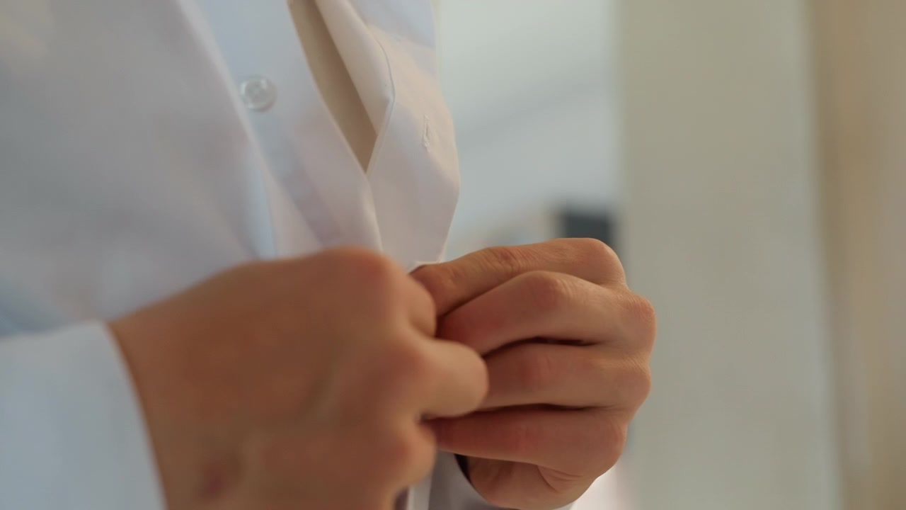 Mani in primo piano che abbottonano una camicia bianca, sposo calmo che si prepara per il matrimonio in una suite illuminata delicatamente, delicate regolazioni dei polsini, dita concentrate, intimo rituale mattutino, attesa e respiro calmo catturati.