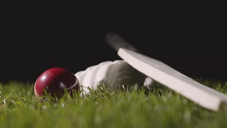 studio cricket still life with close up of ball rolling next to bat and glove lying in grass 2