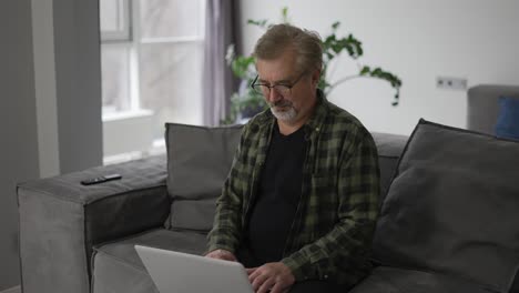 Senior-older-mature-man-typing-on-laptop-browsing-internet-sit-on-sofa-alone