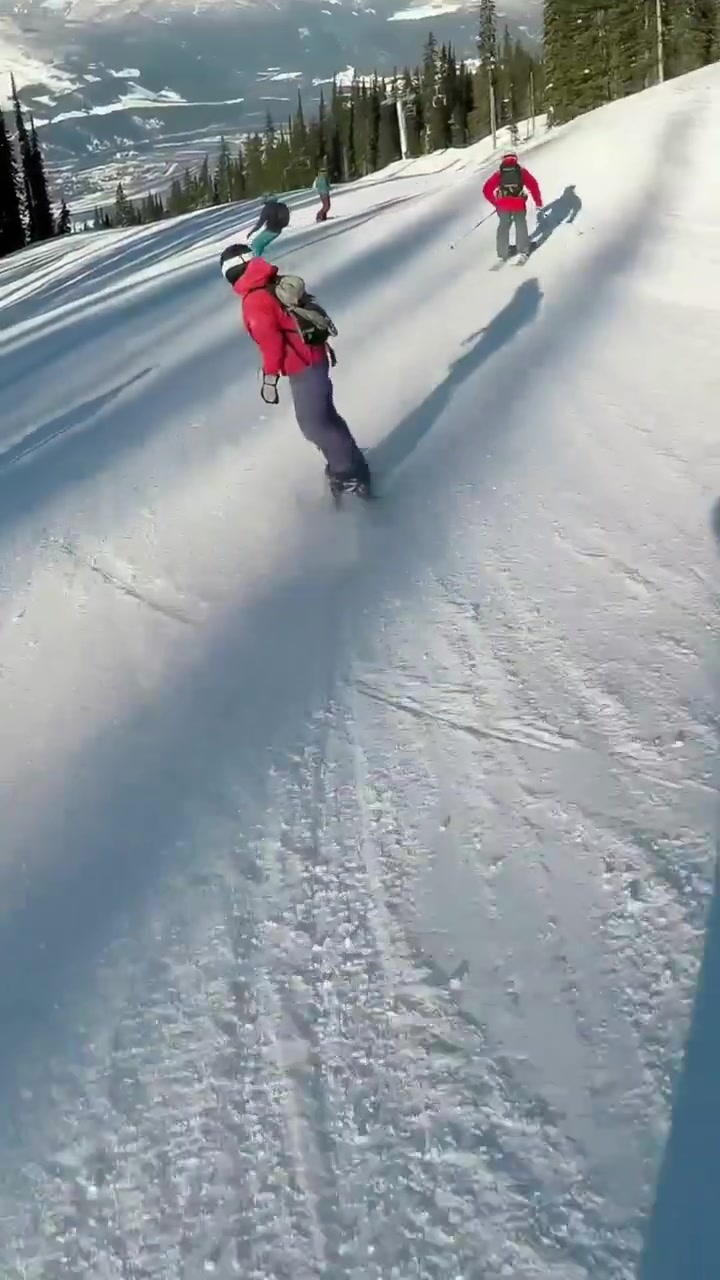 Video verticale: Telecamera che segue uno snowboarder con giacca rossa che scende una pista di montagna con amici, lanciando neve.