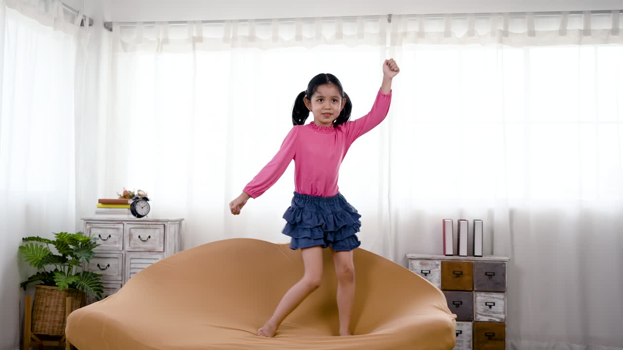 4K 50fps, gadis menggemaskan, bersenang-senang menari di sofa cokelat, dengan gerakan tari yang lucu, dengan wajah bahagia. Konsep keluarga bahagia