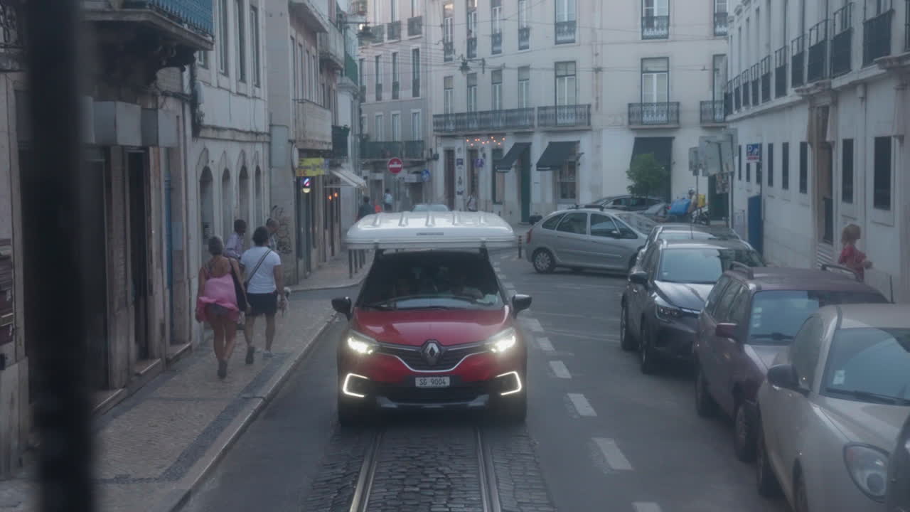 Small red car follows trolley tracks behind an electric trolley as people walk the streets of Lisbon, Portugal, with cars parked along the roadside, capturing the lively urban atmosphere.