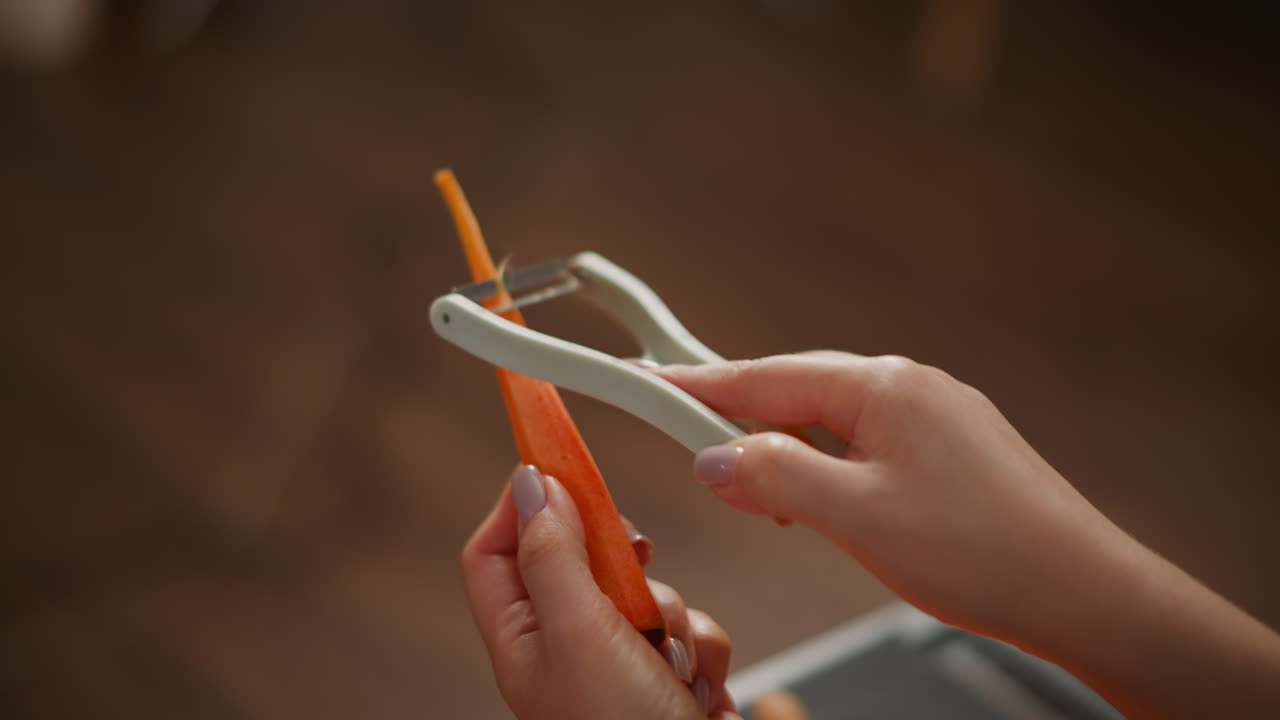 Close up of person peeling pine-shaped carrot with white peeler, neatly manicured hands holding orange vegetable, background features warm toned wooden floor softly blurred in kitchen environment