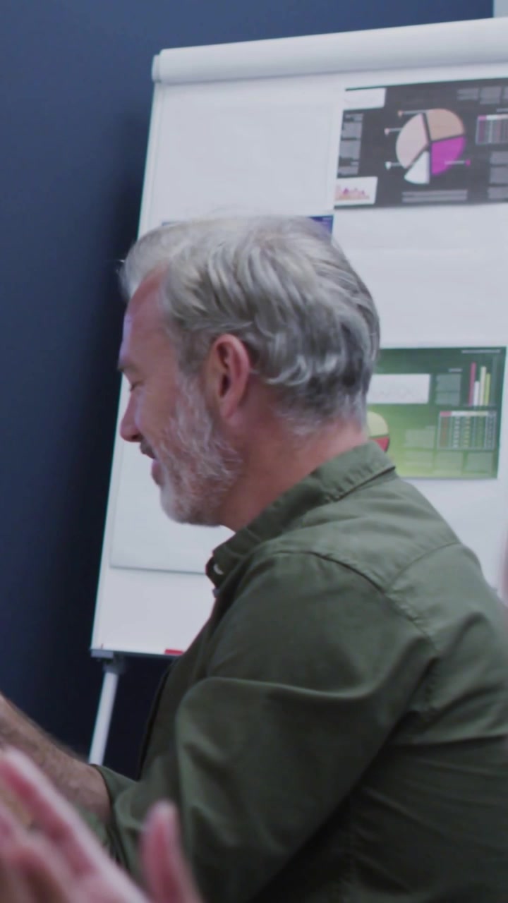 Video verticale: Uomo in una riunione di lavoro che gira la testa dopo un applauso, con numeri che scorrono su un flip chart.