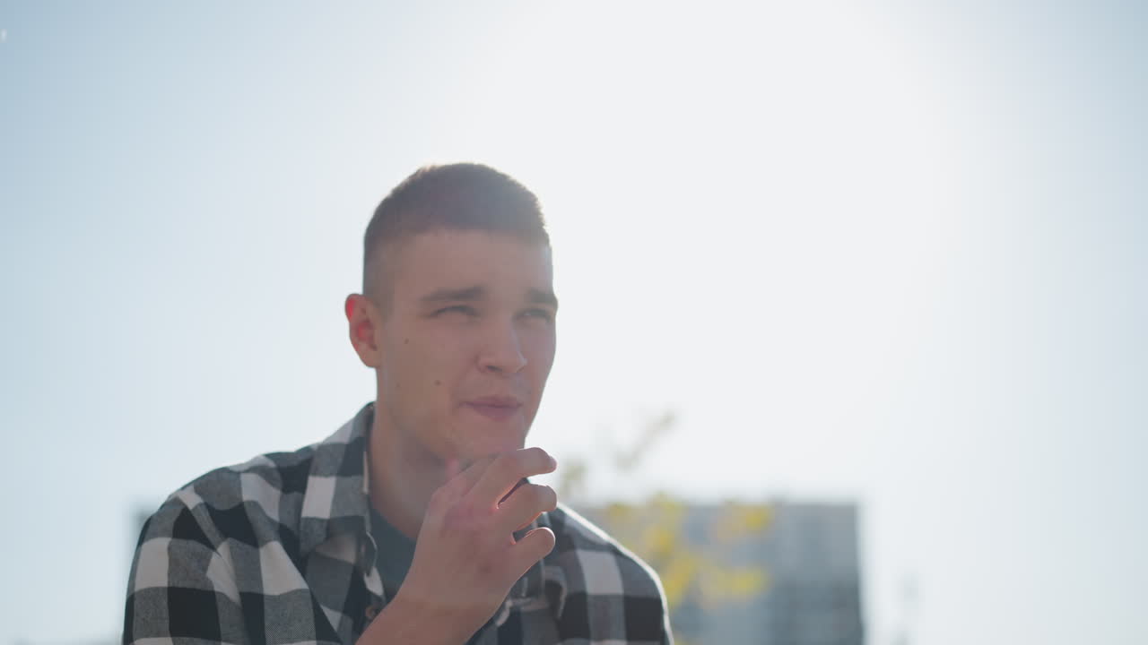 young man outdoors during sunny day puts sweet in mouth while sunlight creates soft glowing effect around him as he gently smiles with blurred buildings and trees in distant background