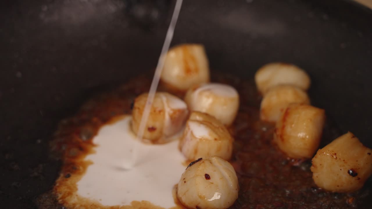 Seafood dish preparation, chef adding three spoons of coconut milk into a shallow frying pan with buttery golden brown pan grilled scallops, close up shot