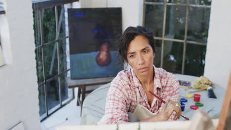 Biracial-female-artist-in-apron-holding-brush-and-looking-at-canvas-in-studio,-slow-motion