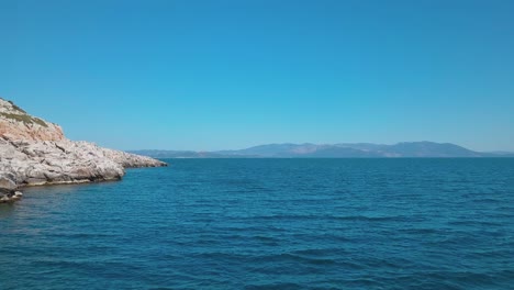 Forward-low-altitude-aerial-shot-next-to-a-rocky-shore-over-the-aegean-sea-with-a-view-of-Euvoia