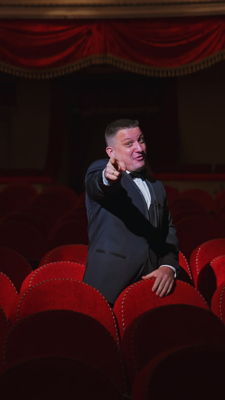Happy man in dark empty theatre hall. Rich man in suit standing among vacant red armchairs in large auditorium and dancing joyfully alone. Vertical video