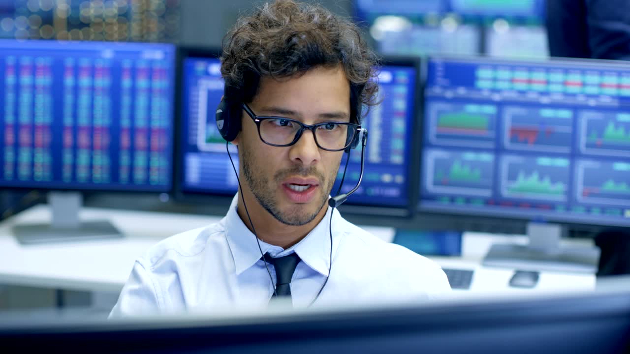 Broker Making Sales with a Headset with His Multi-Ethnic Team of Stock Traders at the Stock Exchange Firm Office is Busy Selling and Buying Bonds and Shares. Displays Show Relevant Graphs and Numbers.