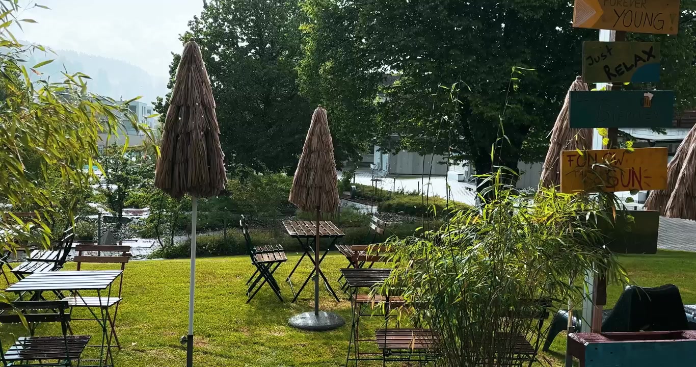 Inquadratura esterna tranquilla di pioggia che cade mentre il sole splende in un verde giardino di ristorante, che mostra tempo estivo, acquazzone di sole, atmosfera fresca, natura, relax e svago di viaggio.
