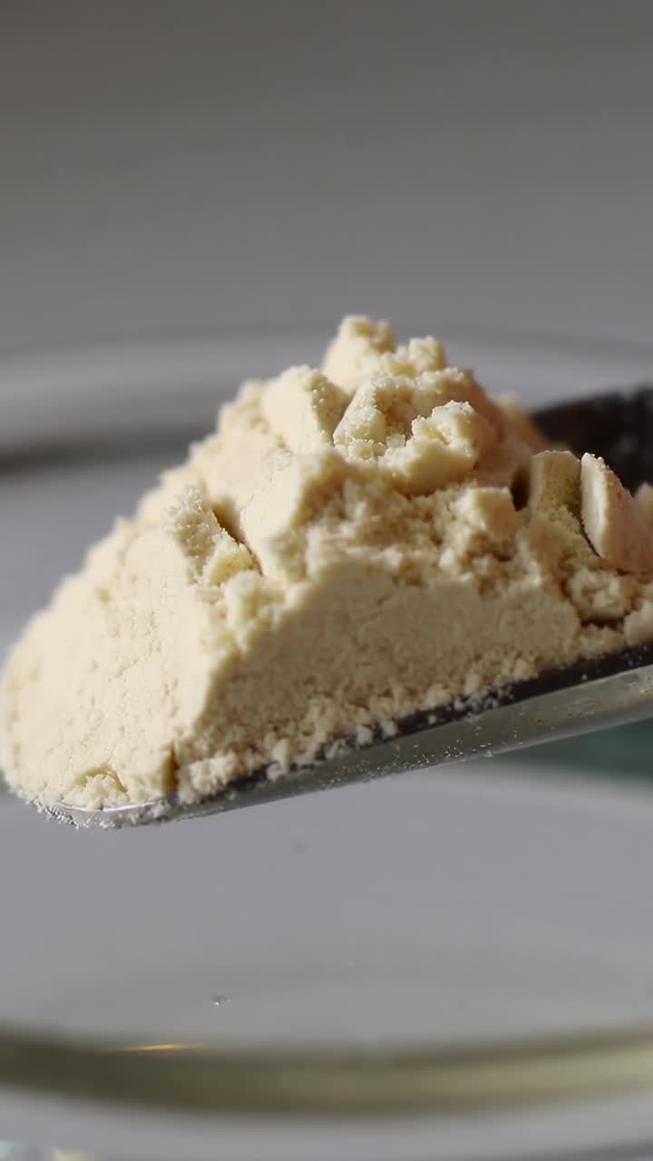 Verticale video: Kantelende metalen lepel met beige poeder boven heldere glazen kom, grijze achtergrond.