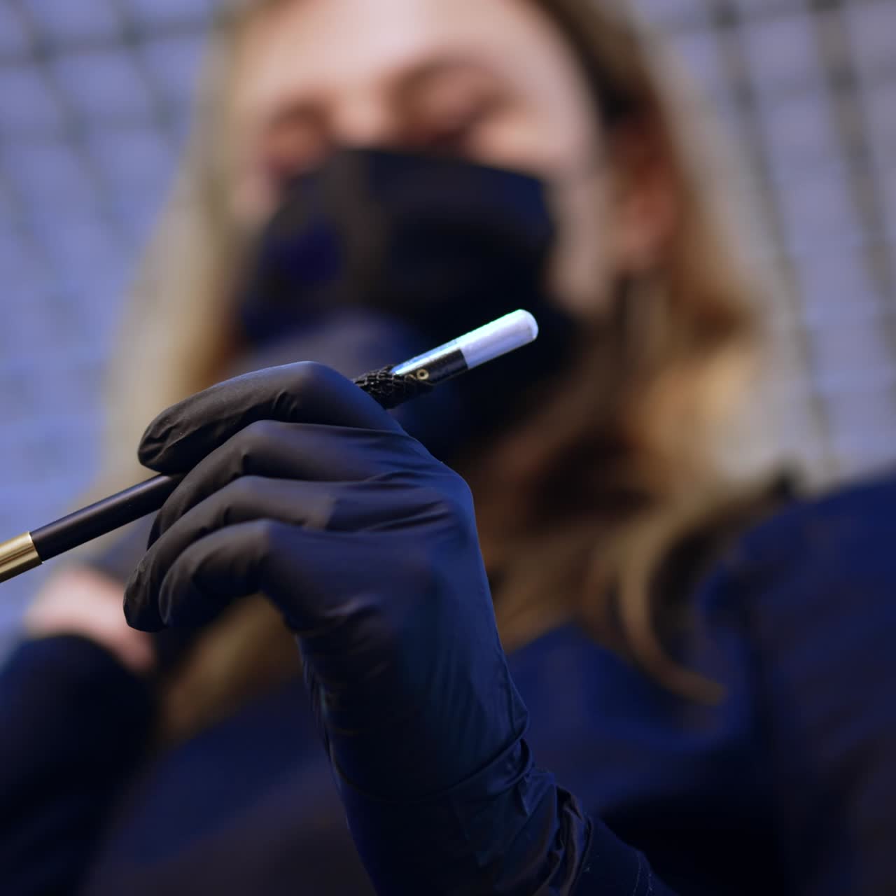 Unrecognized beauty master opens white pencil. Specialist in mask and gloves is going to line the brows for future permanent make up. Low angle view. Blurred backdrop