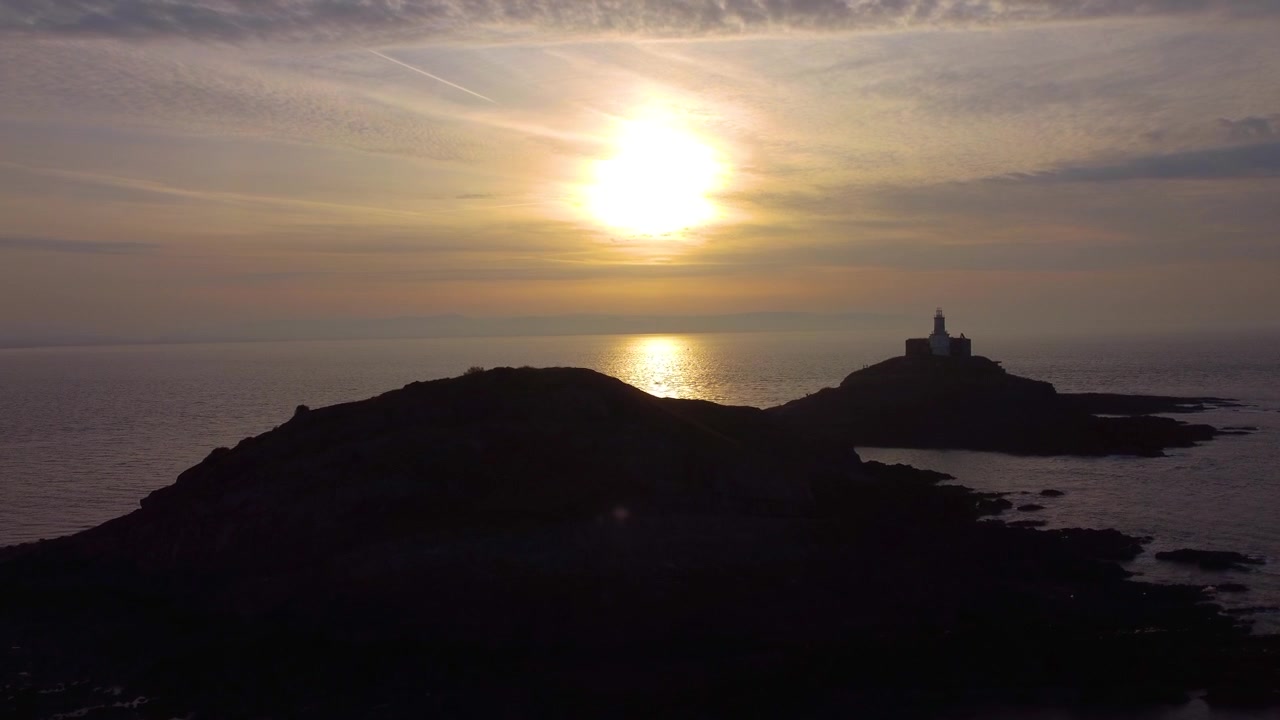 Imagem aérea ascendente do Farol de Mumbles ao nascer do sol. Reflexo dourado do sol no oceano com nuvens dinâmicas. Perfeito para turismo, documentários de viagem. País de Gales, Reino Unido. 4K.