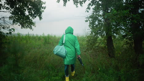 vista trasera de una mujer con un impermeable verde que lleva una bolsa de color menta y un paraguas mientras usa botas de lluvia amarillas, caminando por un camino rodeado de vegetación y árboles