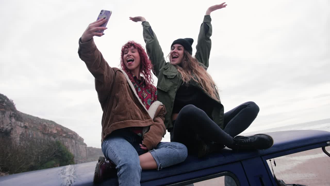 Women sitting on top of van taking selfie with smartphone