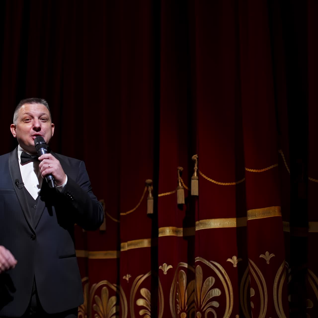Konferencier med mikrofon annoncerer forestilling foran publikum fra scenen. Baggrund med teatergardiner.