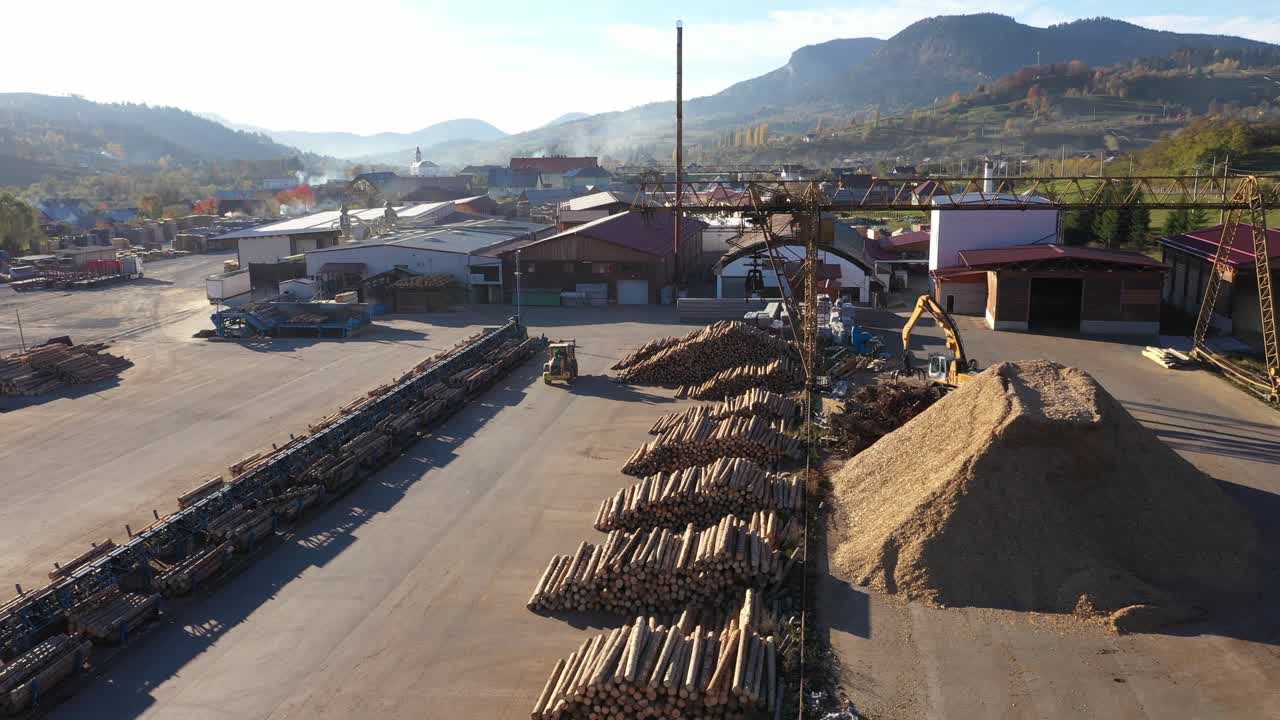 An aerial shot shows an excavator in action at a large sawmill. The heavy machinery works with a massive pile of sawdust, a byproduct of the timber and biomass energy industry