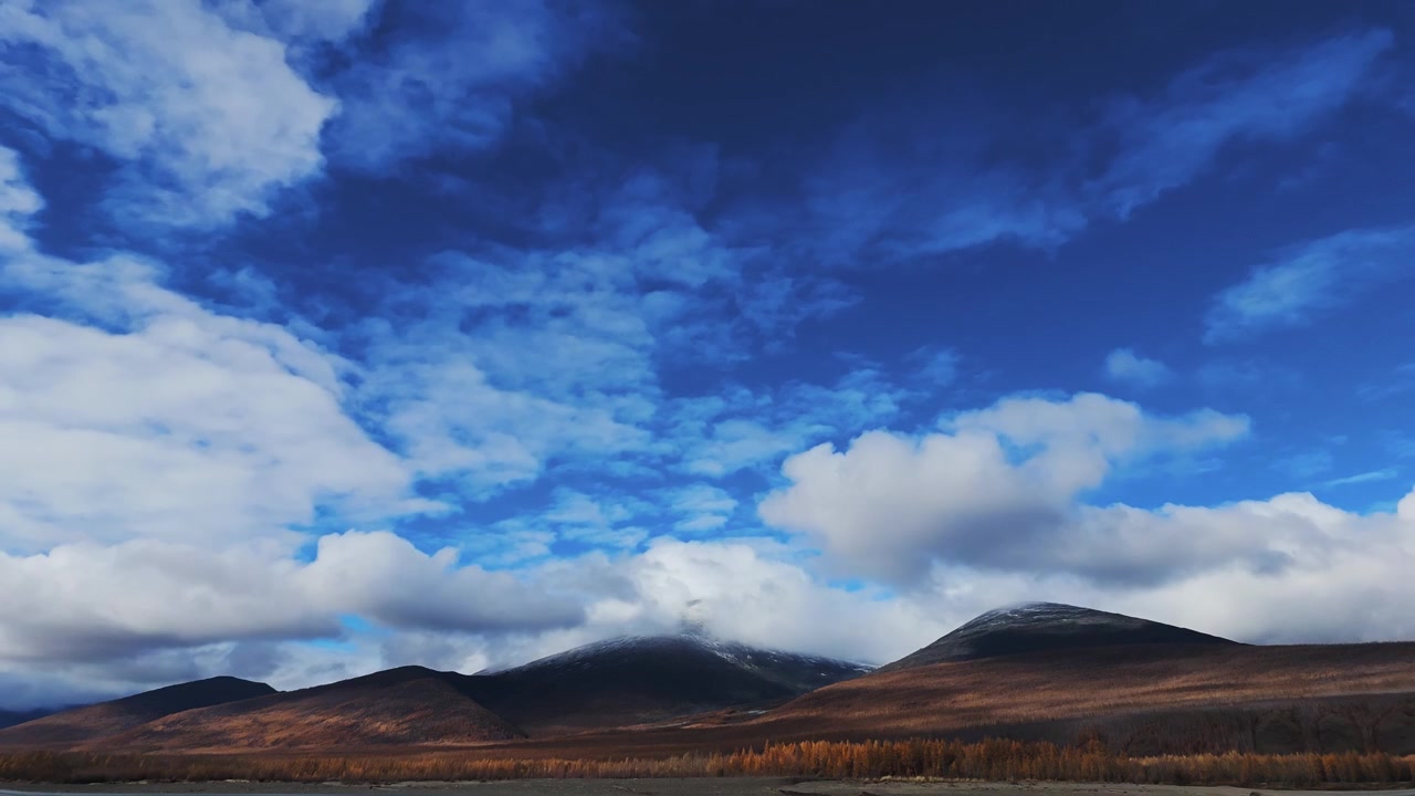 Time-lapse of clouds drifting left to right across a vibrant blue sky above snow-dusted mountain peaks, with golden-brown trees and rolling terrain in the foreground.
