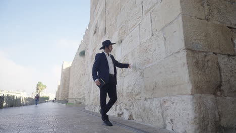 an orthodox man in a black suit and hat walks down an alley by the walls of the old city, also known as the city of david, in jerusalem