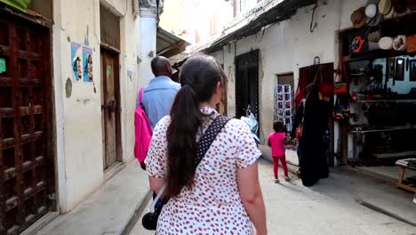 Caucasian-girl-walking-in-street-with-some-locals-and-stores-in-Stone-Town,-Zanzibar