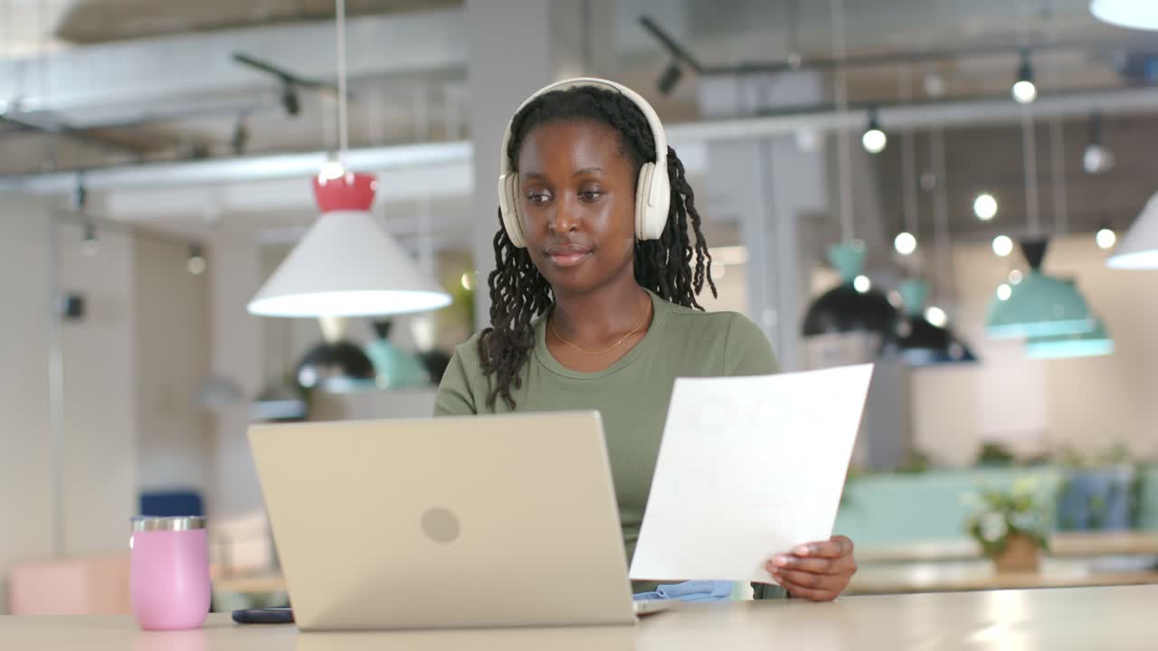 Donna afroamericana seduta alla postazione di lavoro, solleva un foglio e verifica con il laptop, indossando le cuffie.