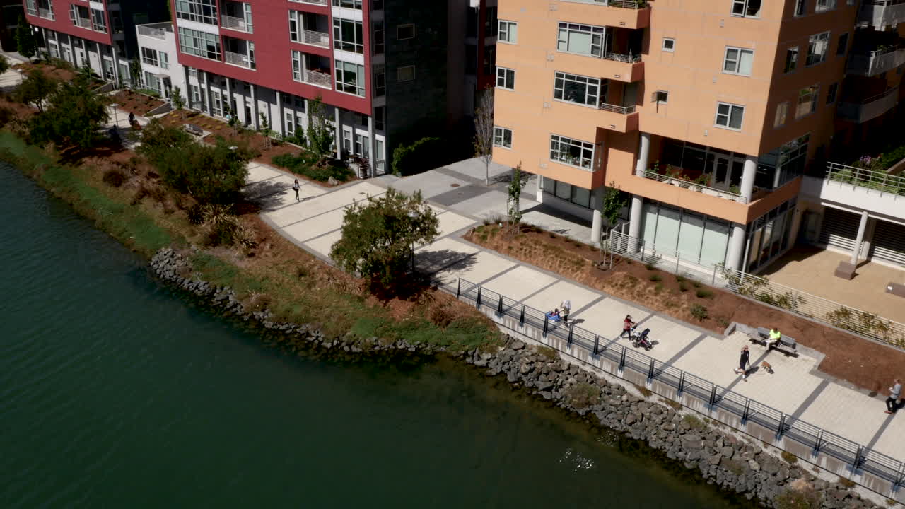 People walk and jog along a waterfront promenade next to modern residential buildings on a sunny day