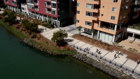 people walk and jog along a waterfront promenade next to modern residential buildings on a sunny day
