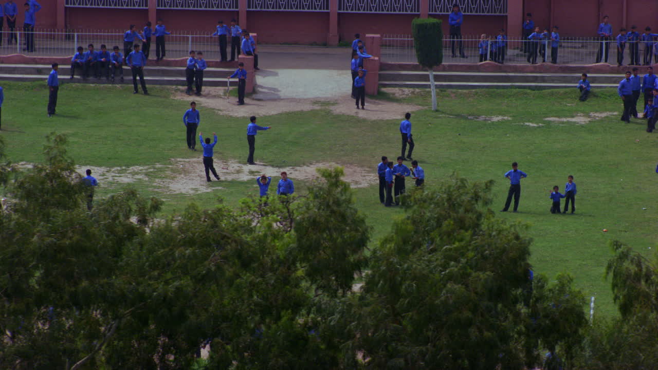 나무를 지나가는 지상 조감도에서 놀고 있는 학교 학생들, 파란색 바지와 셔츠를 입고 있는 학생들, 일부는 땅에 잔디가 없고, 배경에는 학교 건물이 없습니다