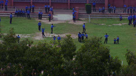 school students playing in the ground aerial view passing the trees, students are in blue pants and shirts, some part is missing of grass in the ground, school building in the background