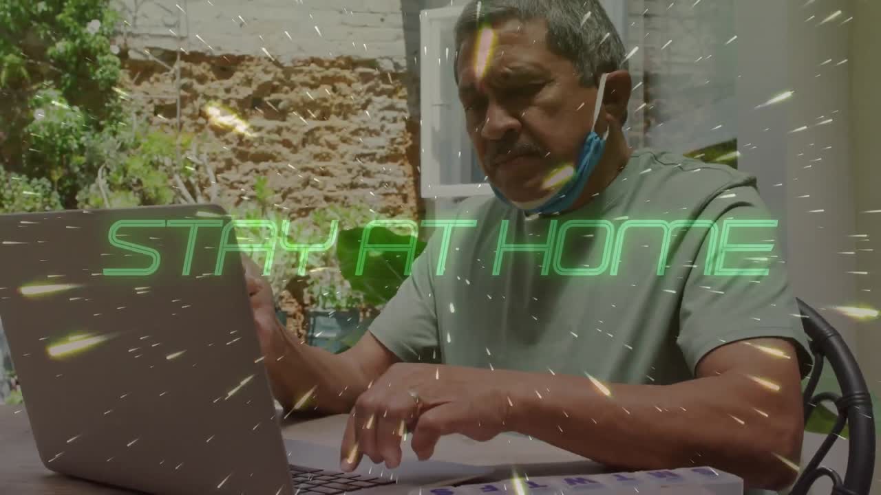 Animation of stay at home text over senior man using laptop wearing face mask