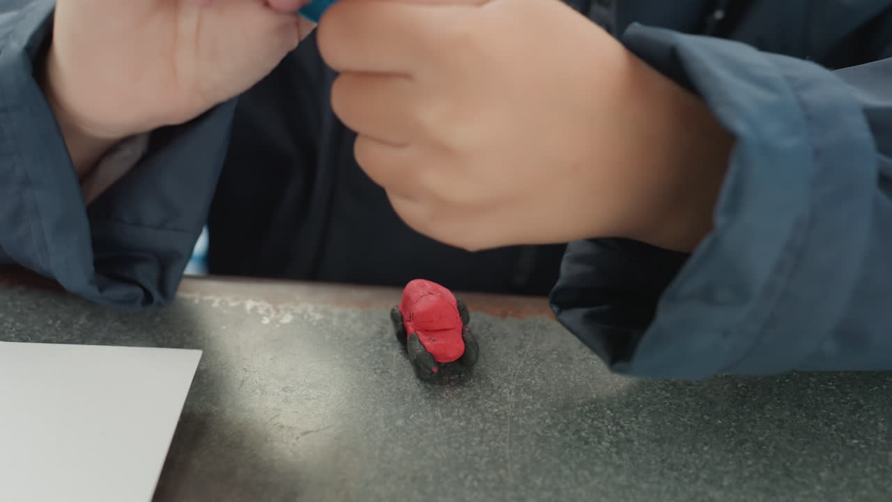 Focused child hands shaping red clay. Outdoor bench craft session showing small eraser on metal desk beside paper and pencil, closeup on fingers molding blue tool, slow methodical motion, calm park