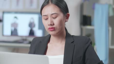 비즈니스 슈트를 입은 아시아 여성 디자이너가 노트북에서 일하고 스튜디오에서 옷을 디자인하는 동안 생각하고 있습니다.