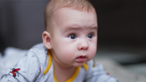 calm peaceful baby boy lies on his belly and looks sideways. curious toddler face portrait close up. grey blurred backdrop.