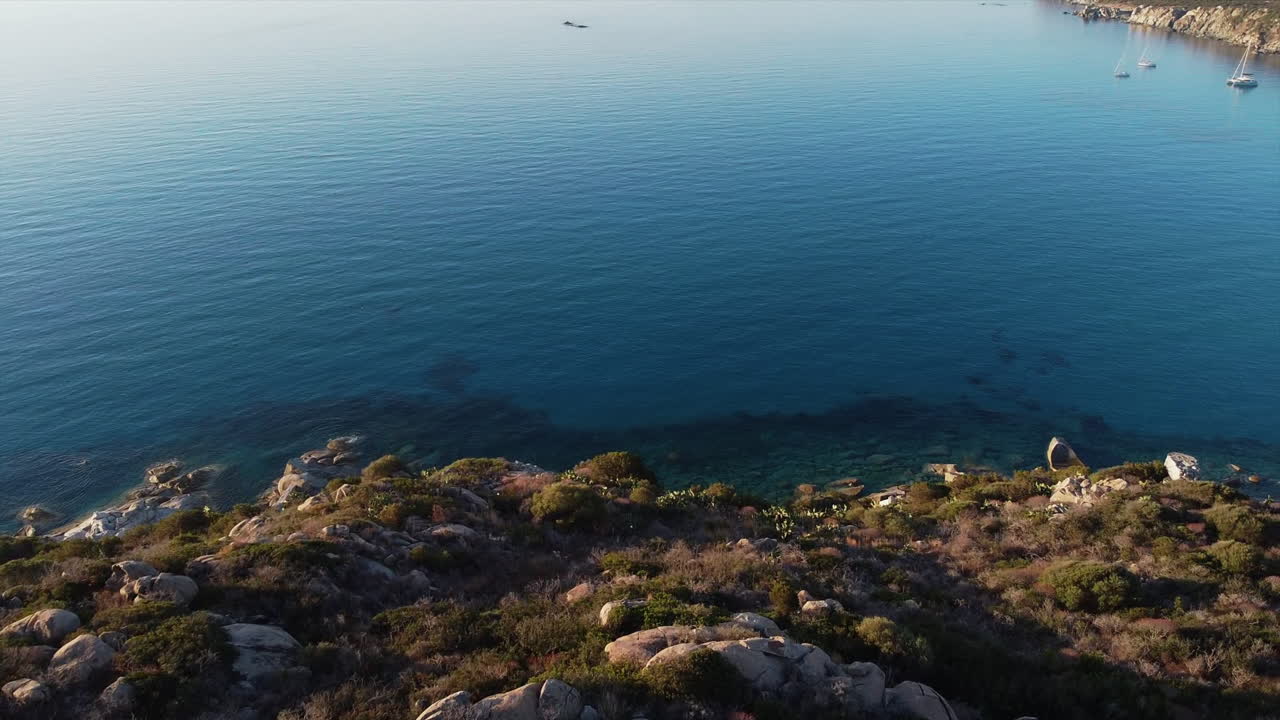 Drone view of cliff with trees and shrubs overlooking clear and blue sea with yacht sailing with silent and empty shore in Italy during bright day