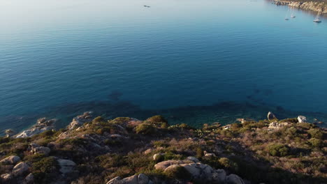 Drone-view-of-cliff-with-trees-and-shrubs-overlooking-clear-and-blue-sea-with-yacht-sailing-with-silent-and-empty-shore-in-Italy-during-bright-day