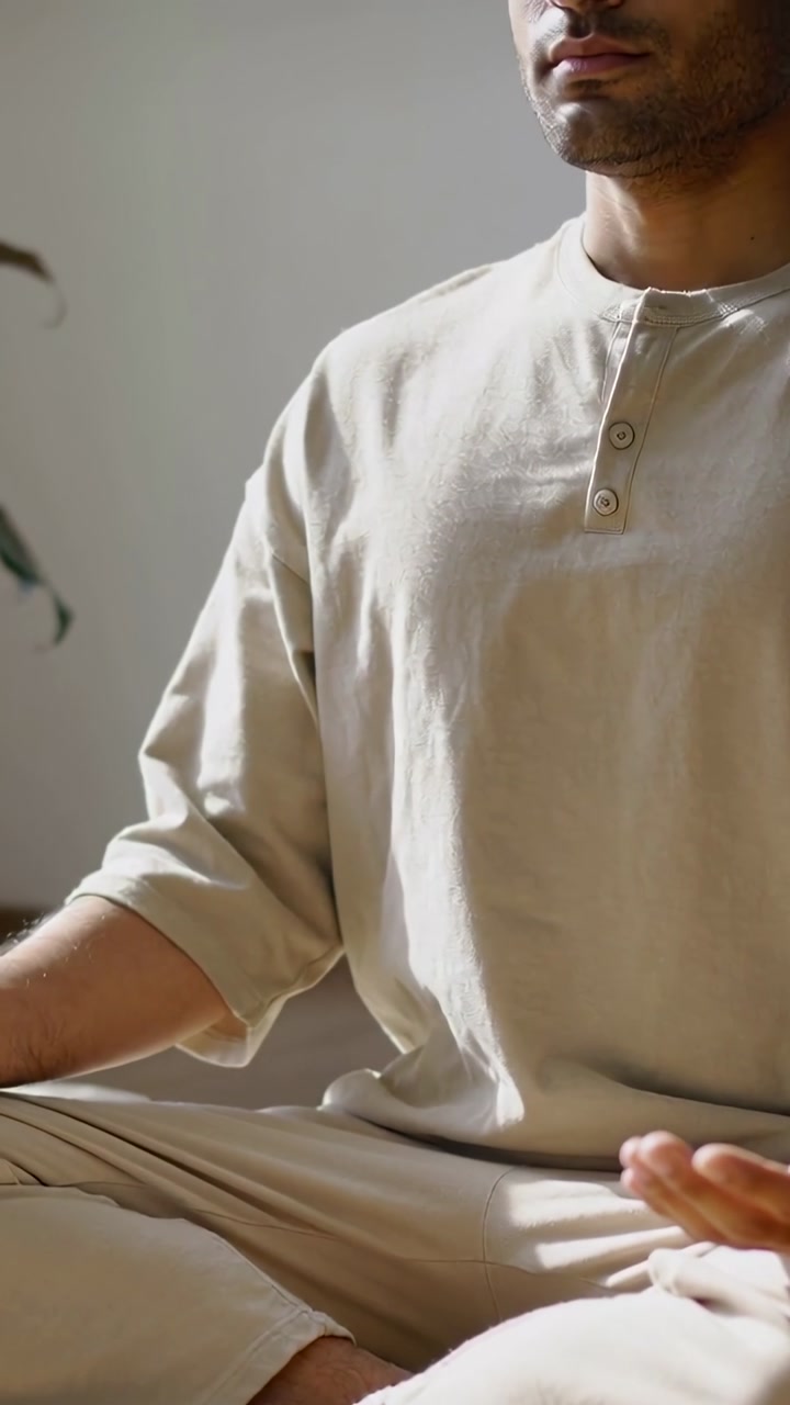 Vertical video: Settling man in beige loungewear moving into meditation on floor, with potted plant