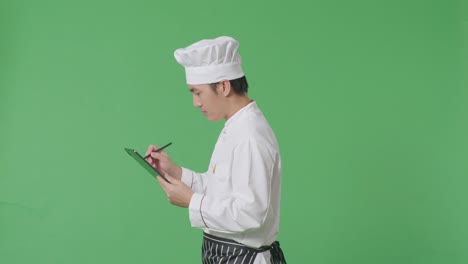 vista lateral del chef asiático tomando nota en una tableta mientras camina en el estudio de fondo de la pantalla verde
