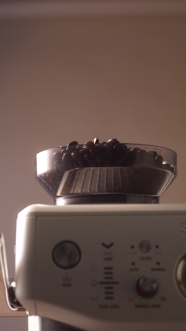 Primo piano al rallentatore di chicchi di caffè tostati che si versano da un contenitore nella tramoggia trasparente di un macinacaffè per espresso domestico con portafiltro. Lo sfondo neutro e caldo e la luce soffusa ne esaltano la texture