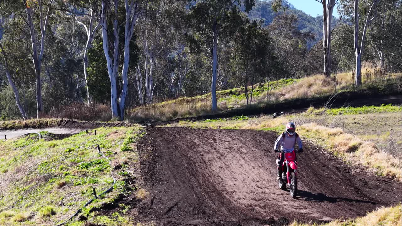 A young rider on a dirt bike accelerates up a dirt ramp, launches into the air, and lands smoothly on a sunlit outdoor motocross track surrounded by eucalyptus trees