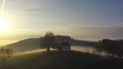 Aerial view of a lone church on a hill in Lembach, Upper Austria, at sunrise. Soft light illuminates the scene as the Mühlviertel is enveloped in mist, creating a peaceful, ethereal atmosphere.